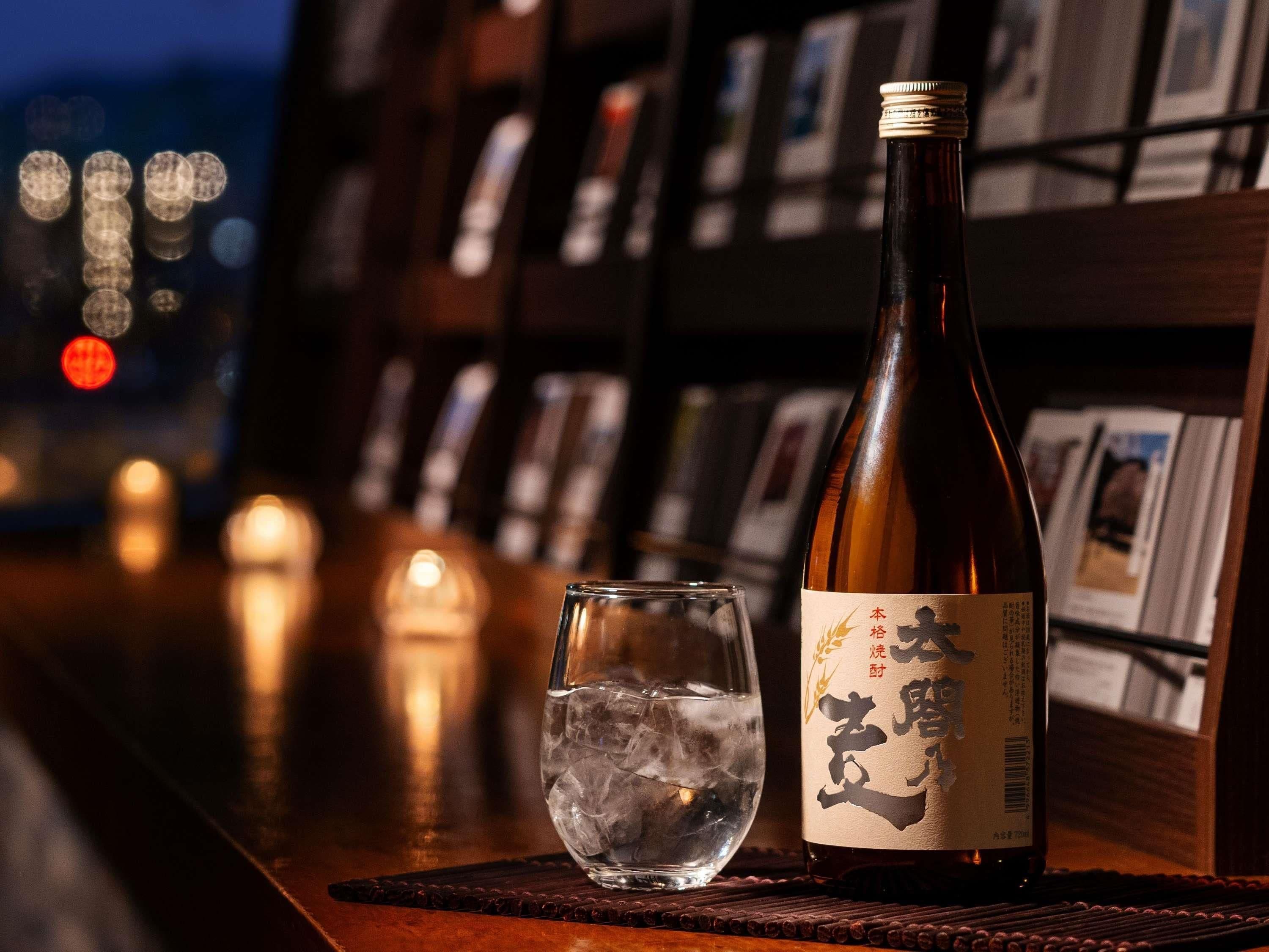 Eine Sake-Flasche und ein Glas mit Eis auf einer Bar, im Hintergrund verschwommen Lichter und Bücherregale.