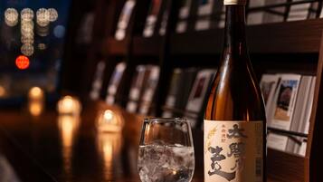Eine Sake-Flasche und ein Glas mit Eis auf einer Bar, im Hintergrund verschwommen Lichter und Bücherregale.