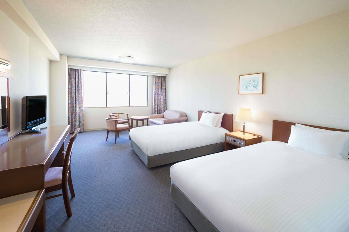 Une chambre d’hôtel avec deux lits, un bureau avec une télévision, une chaise, une table de chevet avec une lampe et une fenêtre avec des rideaux.