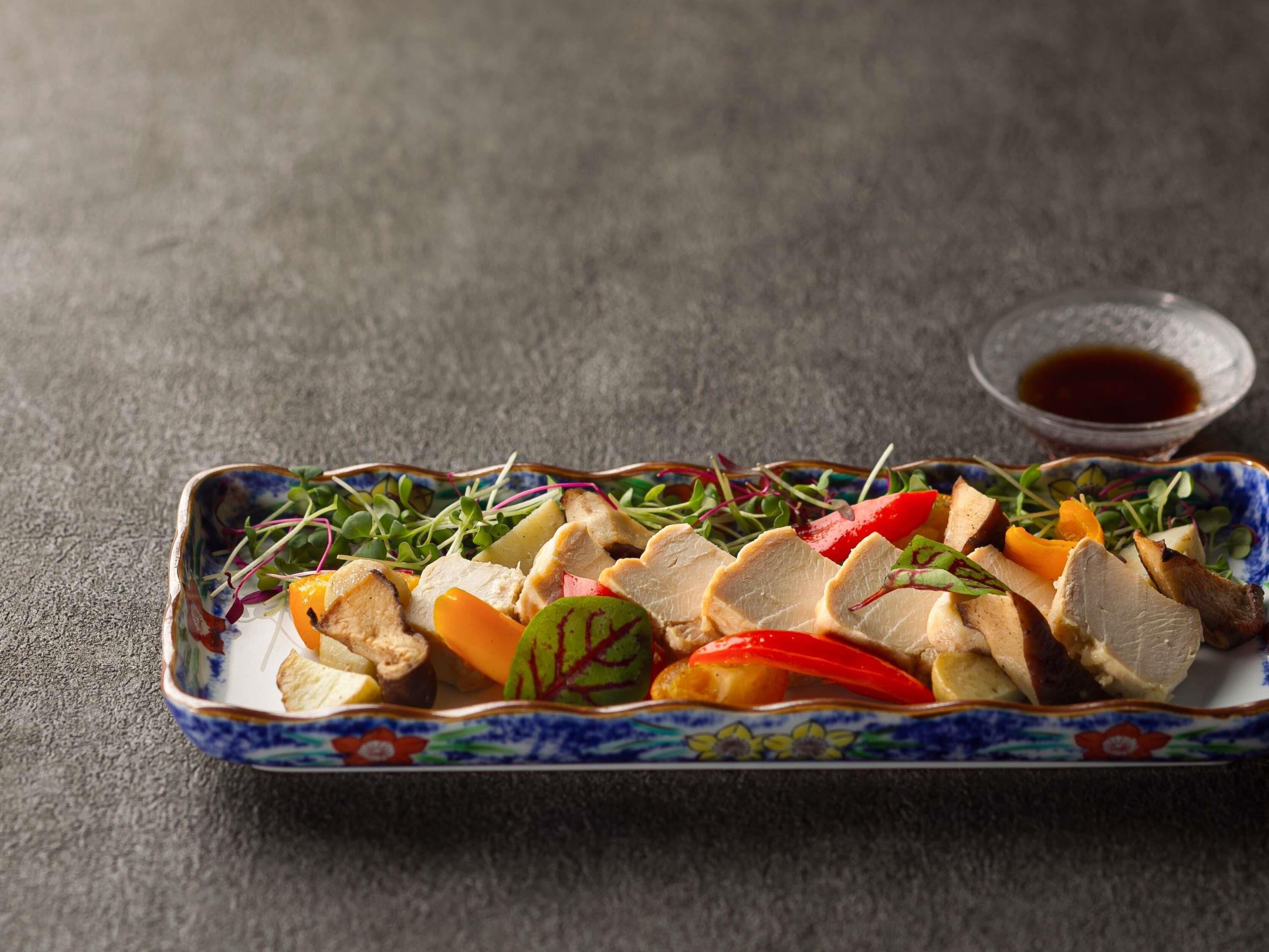 Une assiette rectangulaire ornée d’un motif floral, contenant des morceaux de poulet, des légumes et un petit bol de sauce.