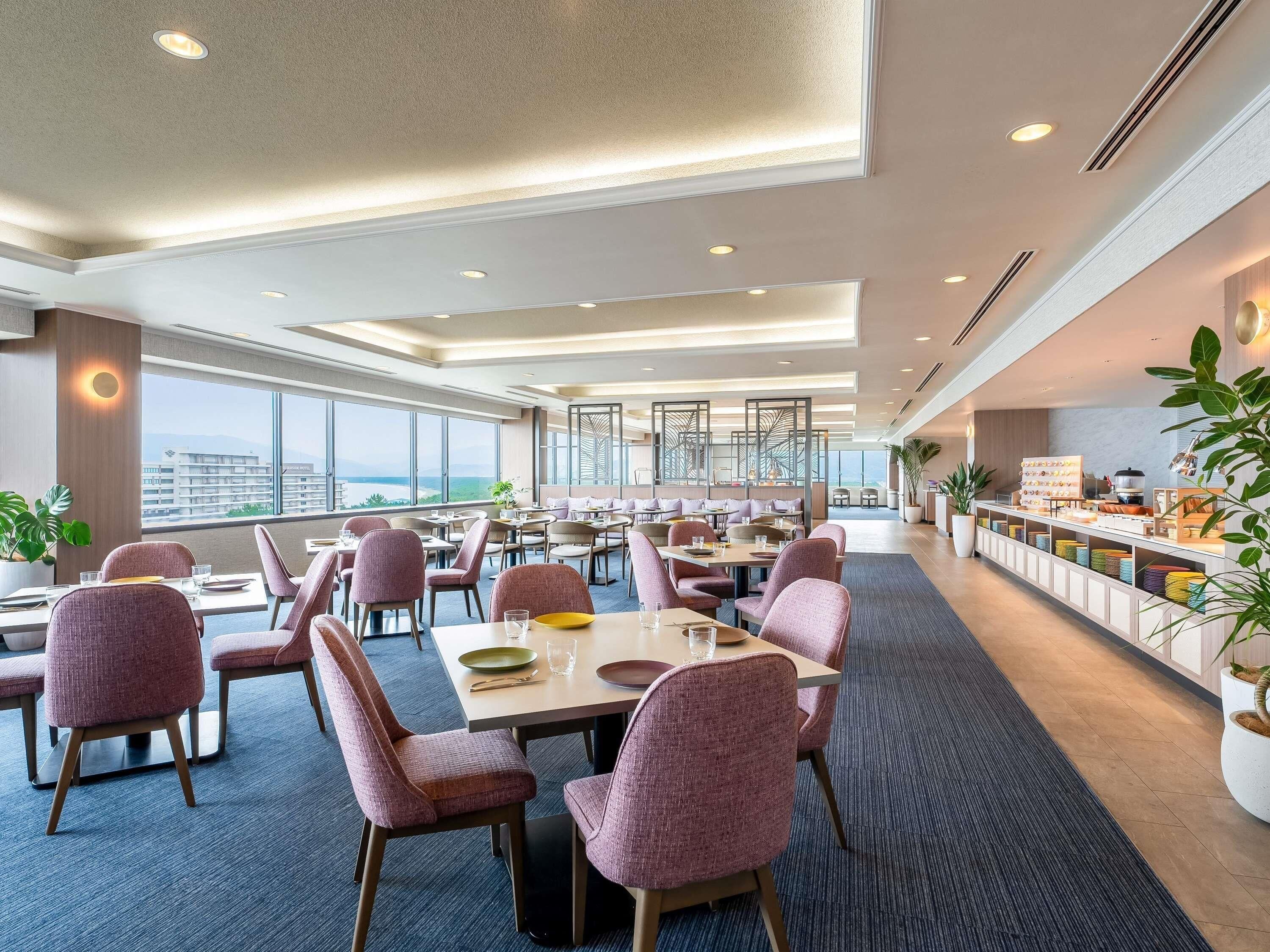 Comedor de hotel con varias mesas y sillas, una barra de buffet y ventanas amplias con vistas.