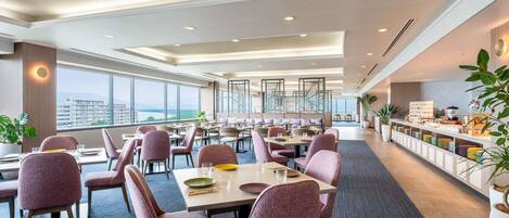Comedor de hotel con varias mesas y sillas, una barra de buffet y ventanas amplias con vistas.