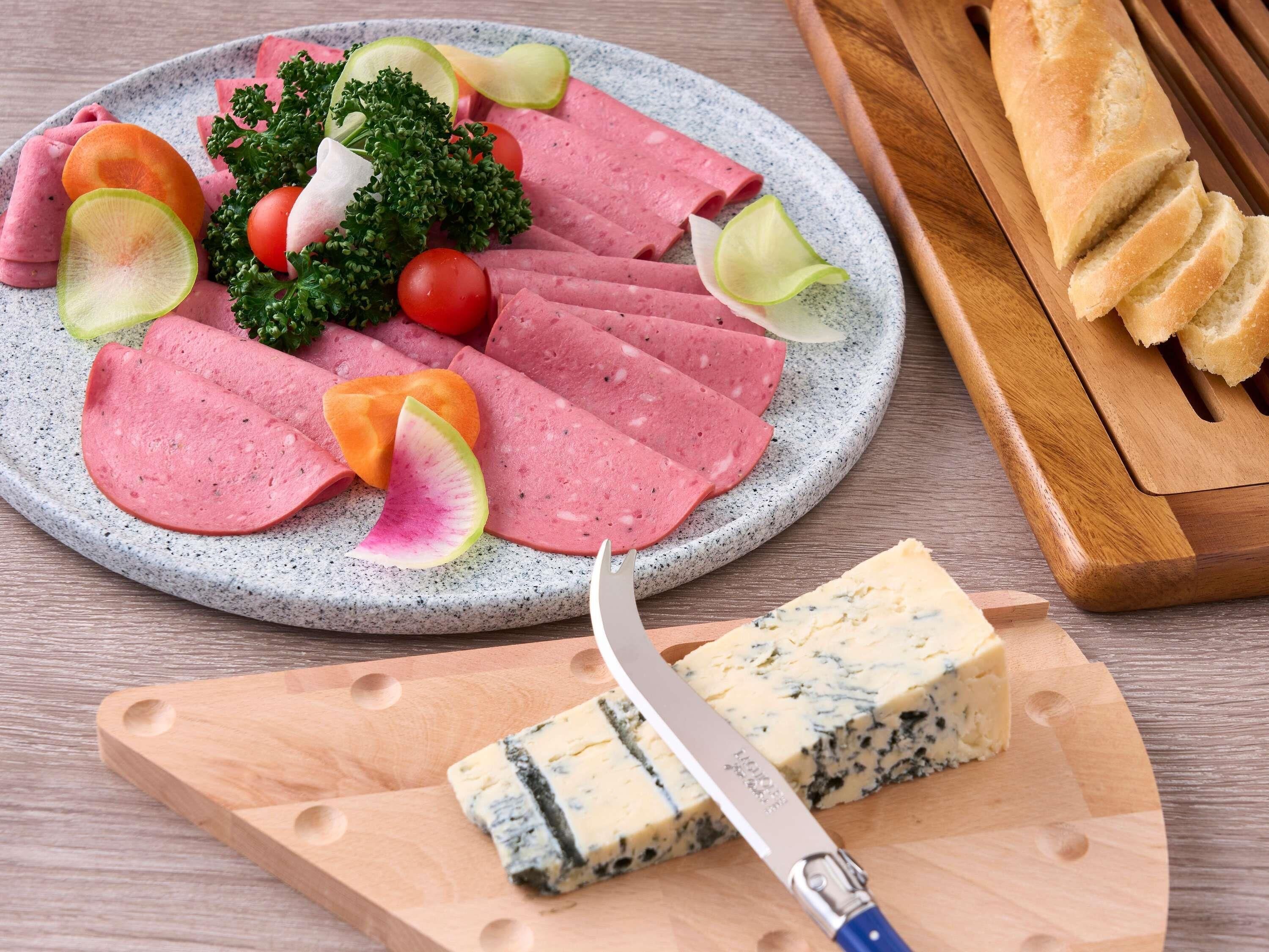 A plate of sliced meats, cheese, and vegetables on a speckled plate, with a knife and a wooden cutting board with cheese on it.