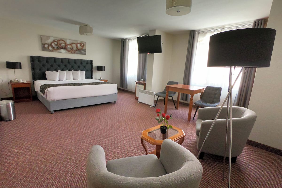 Et hotelværelse med en seng, et skrivebord, to stole, et lille bord med en vase og et fjernsyn på væggen.