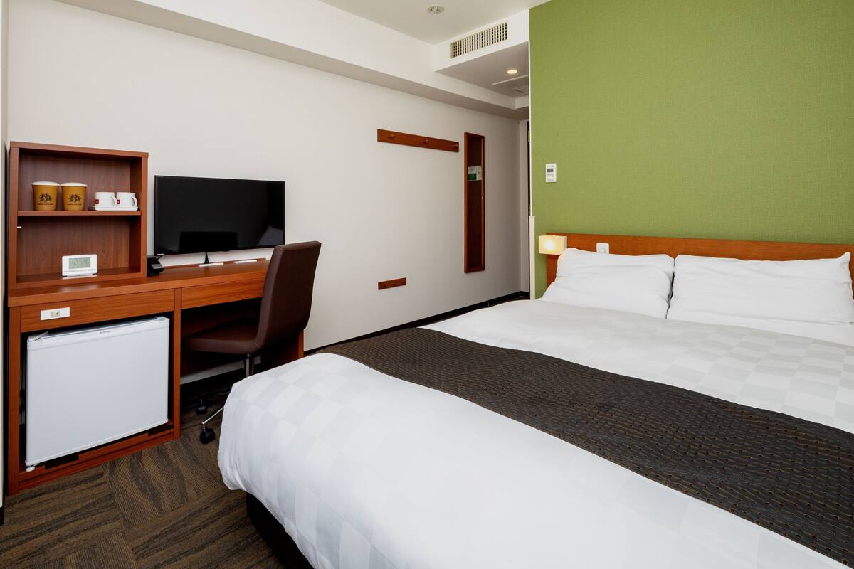 A hotel room with a bed, a desk with a television, a mini-fridge, and a chair.