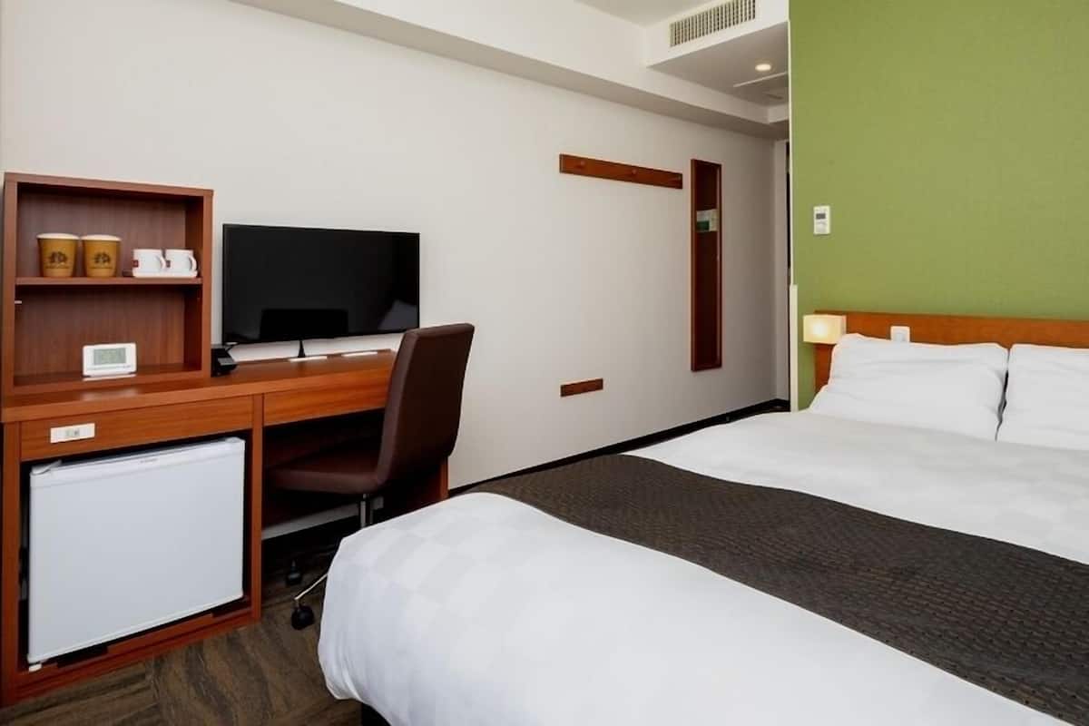 A hotel room with a bed, a desk with a television, a chair, a small fridge, and a green accent wall.