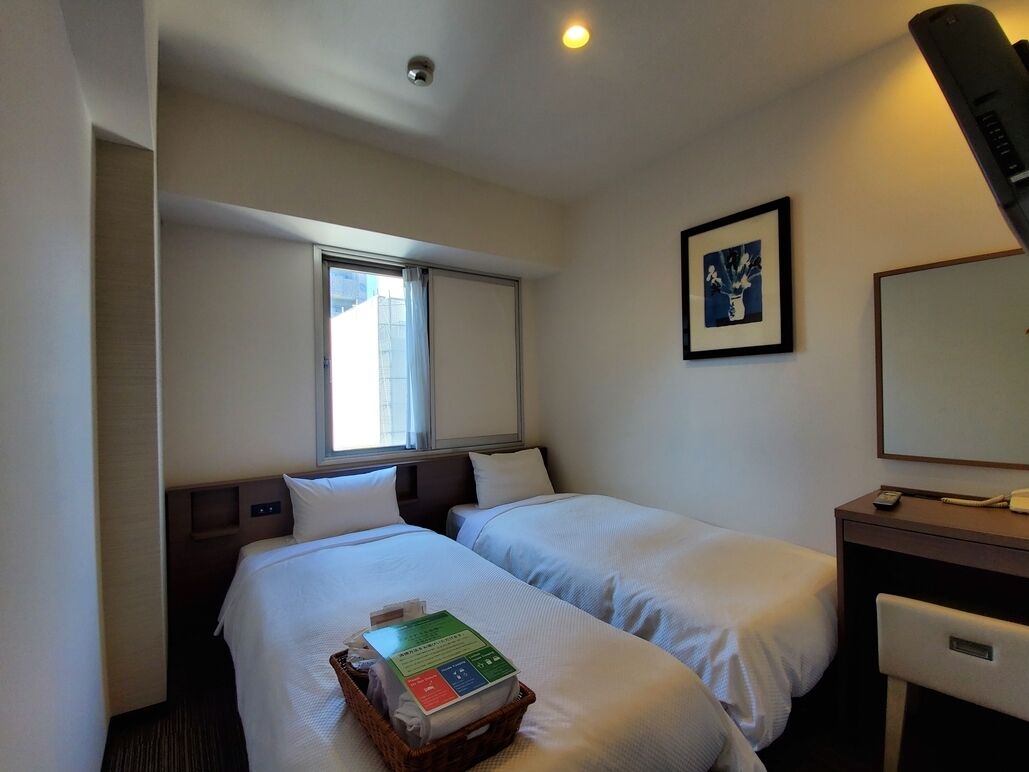 Ein Hotelzimmer mit zwei Betten, einem Schreibtisch, einem Stuhl, einem Fenster und einem gerahmten Bild an der Wand.