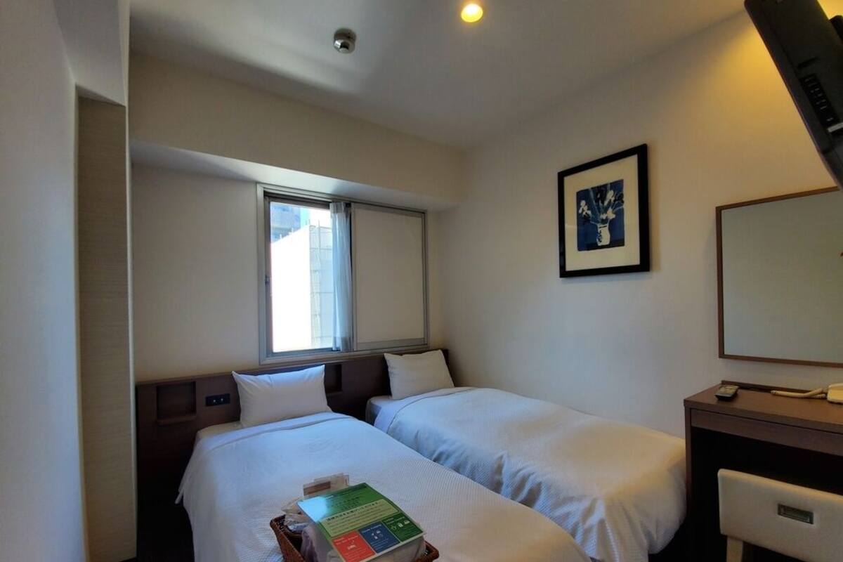 A hotel room with two beds, a desk, a chair, a window, and a framed picture on the wall.
