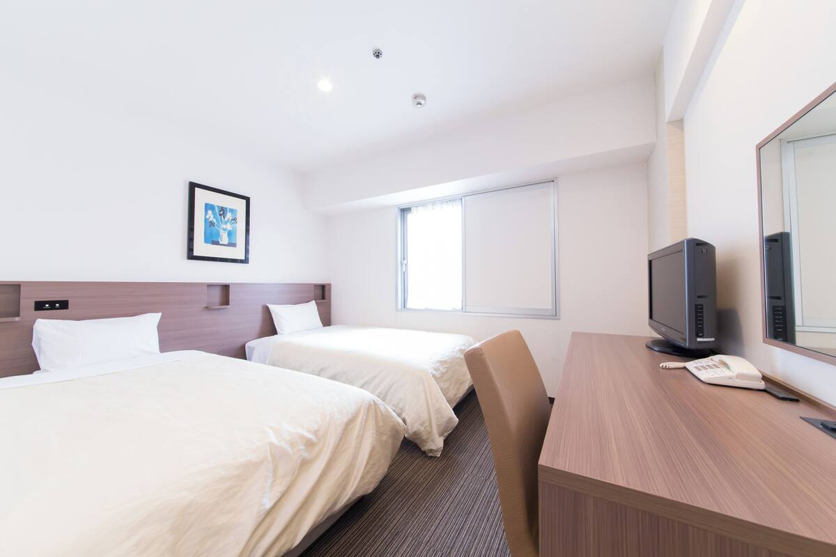 A hotel room with two beds, a desk with a computer and phone, a large window, and a picture on the wall.