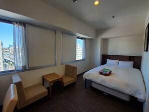 Habitación de hotel con cama, una mesita, dos sillas y vista a la ciudad a través de amplios ventanales.