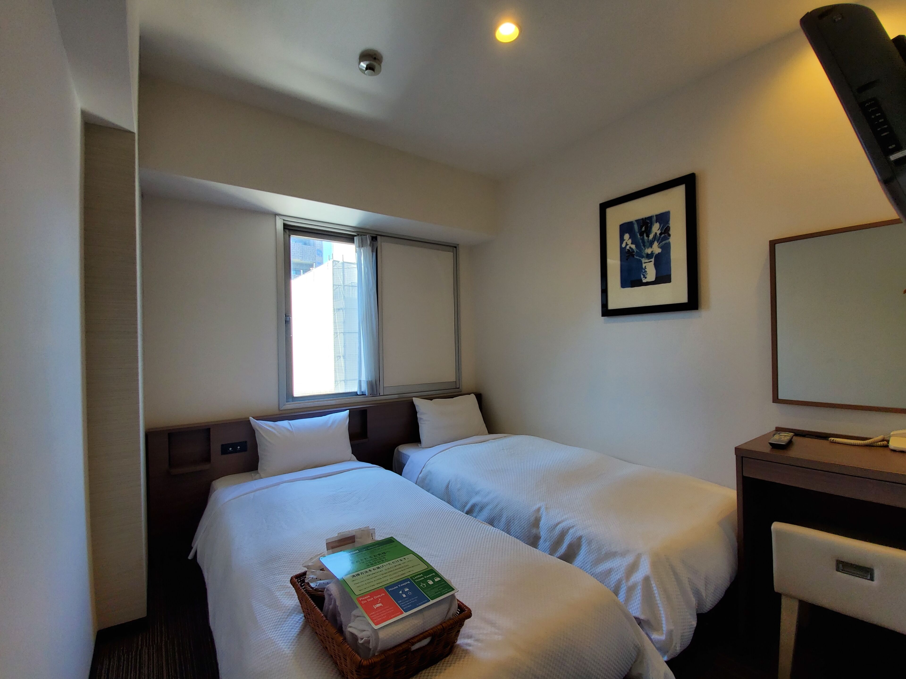 Ein Hotelzimmer mit zwei Betten, einem Schreibtisch, einem Stuhl, einem Fenster mit Vorhängen, einem gerahmten Bild an der Wand und einem Korb mit Gegenständen auf dem Bett.