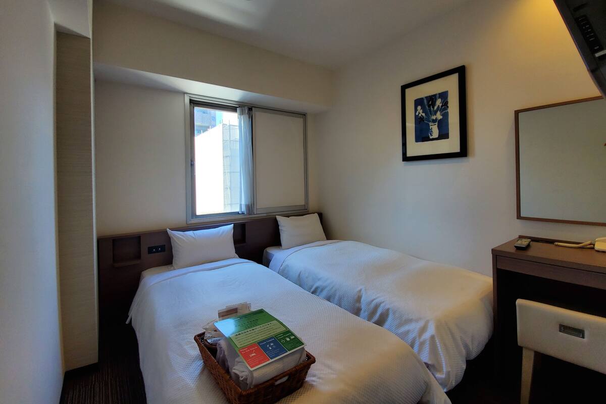 A hotel room with two beds, a desk, a chair, a window with curtains, a framed picture on the wall, and a basket with items on the bed.