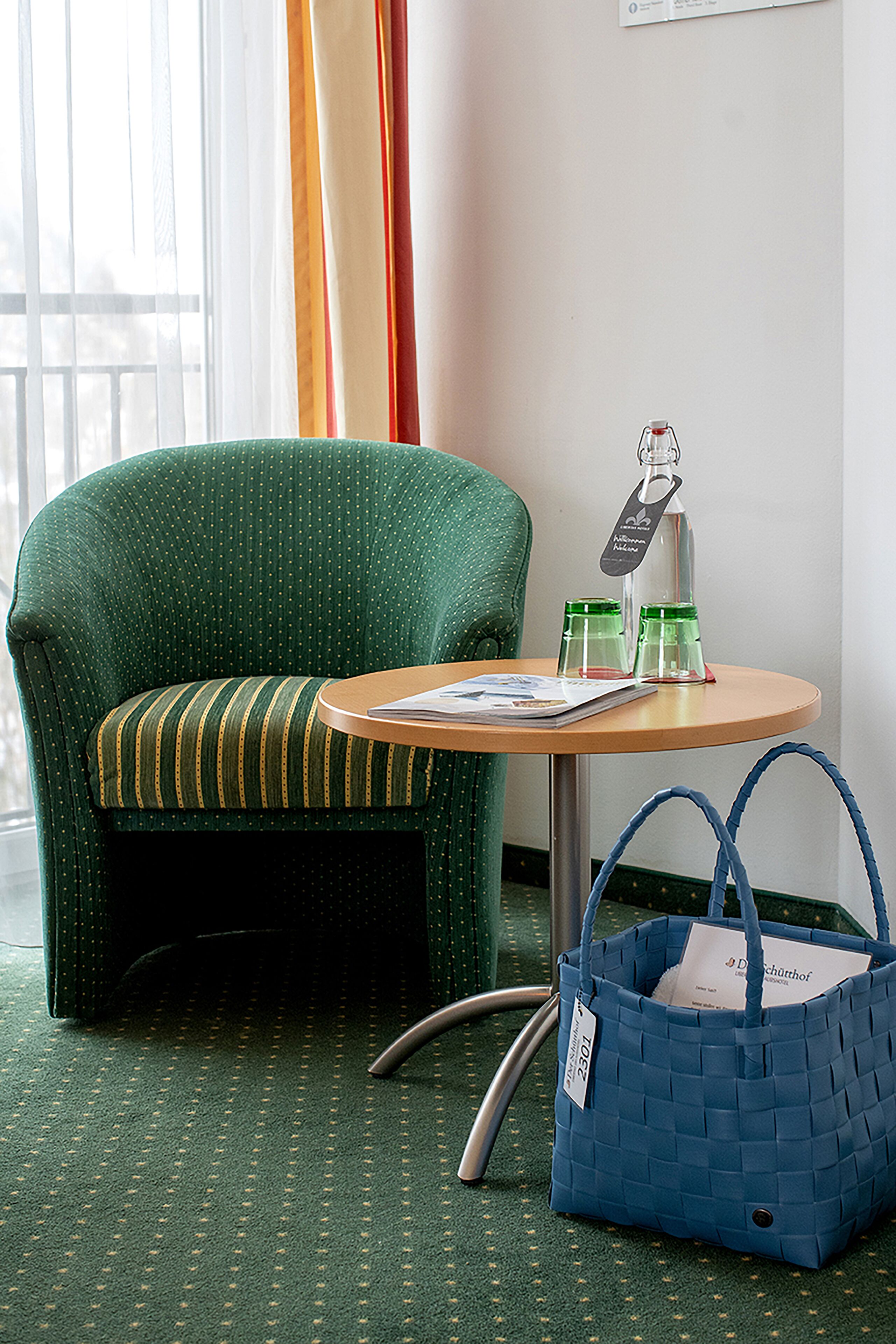 A green upholstered chair with a striped cushion, a small round table with a magazine, two glass bottles, and a blue woven bag with a tag.