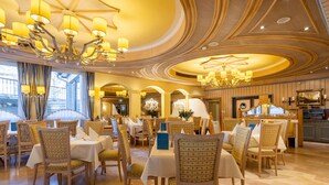 A dining area with multiple tables and chairs, chandeliers, and a kitchen in the background. - Hotel Der Schütthof (Zell am See)