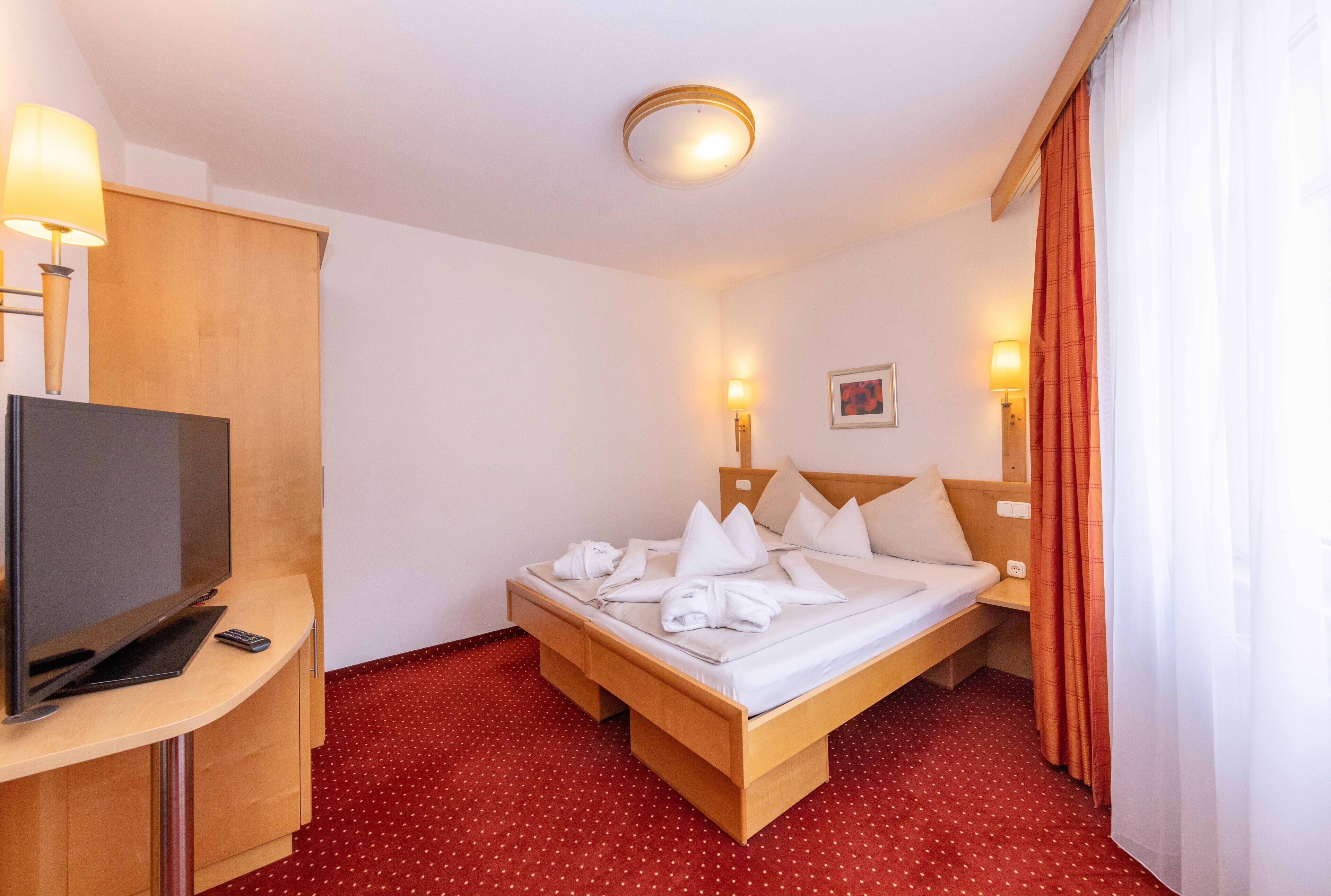 Quarto de hotel com cama, televisão, escrivaninha e tapete vermelho com bolinhas.