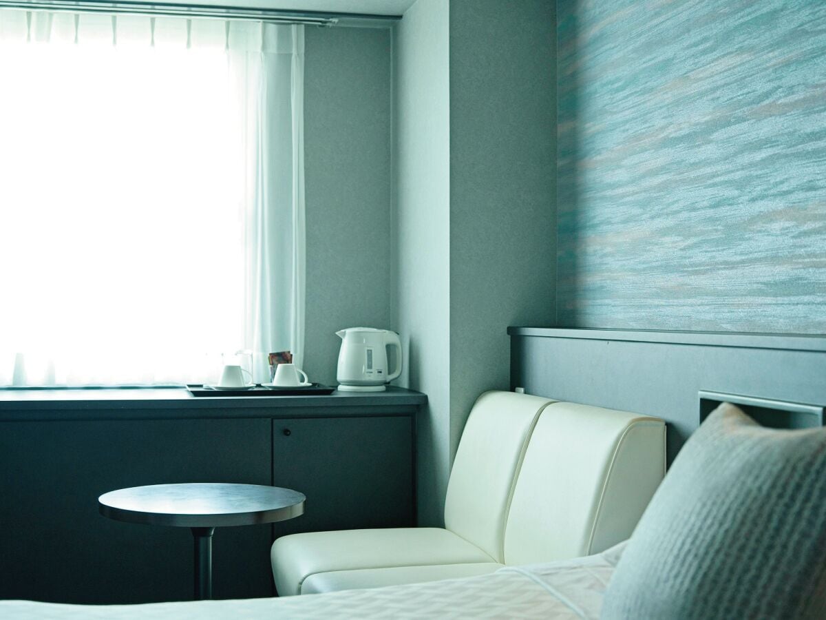 Habitación de hotel con cama, una mesita redonda, ventana con cortinas, radiador fijado a la pared y pared con textura.