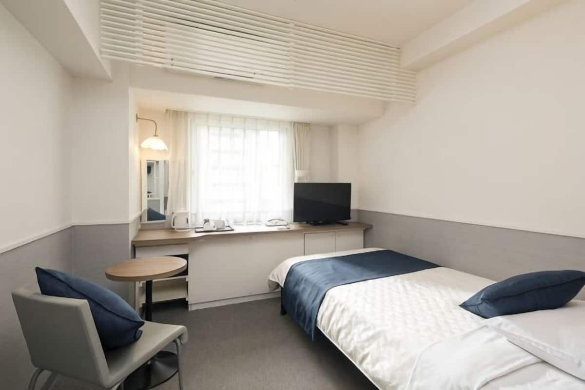 A hotel room with a bed, a desk with a TV, a chair, and a window with curtains.
