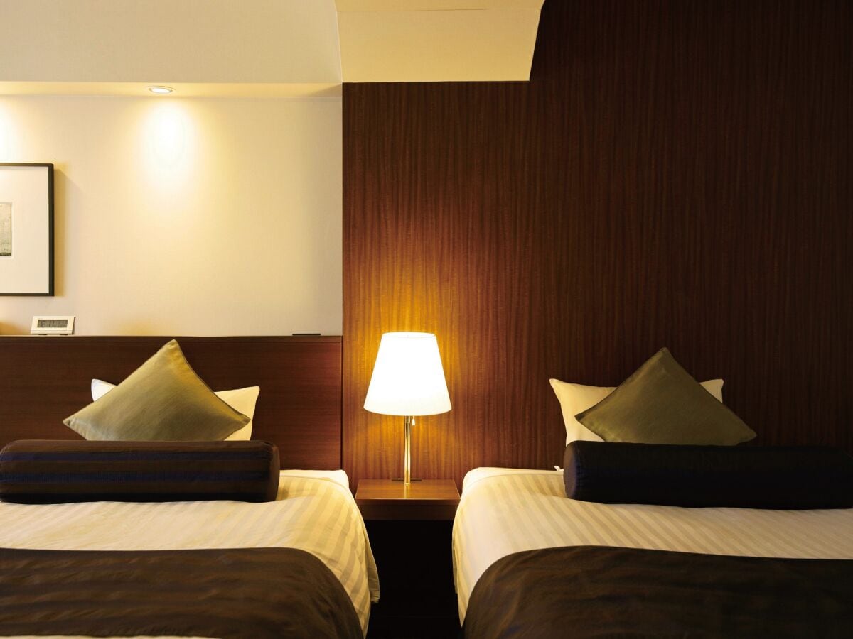 Una camera d'albergo con due letti, un comodino con una lampada e una testiera in legno.