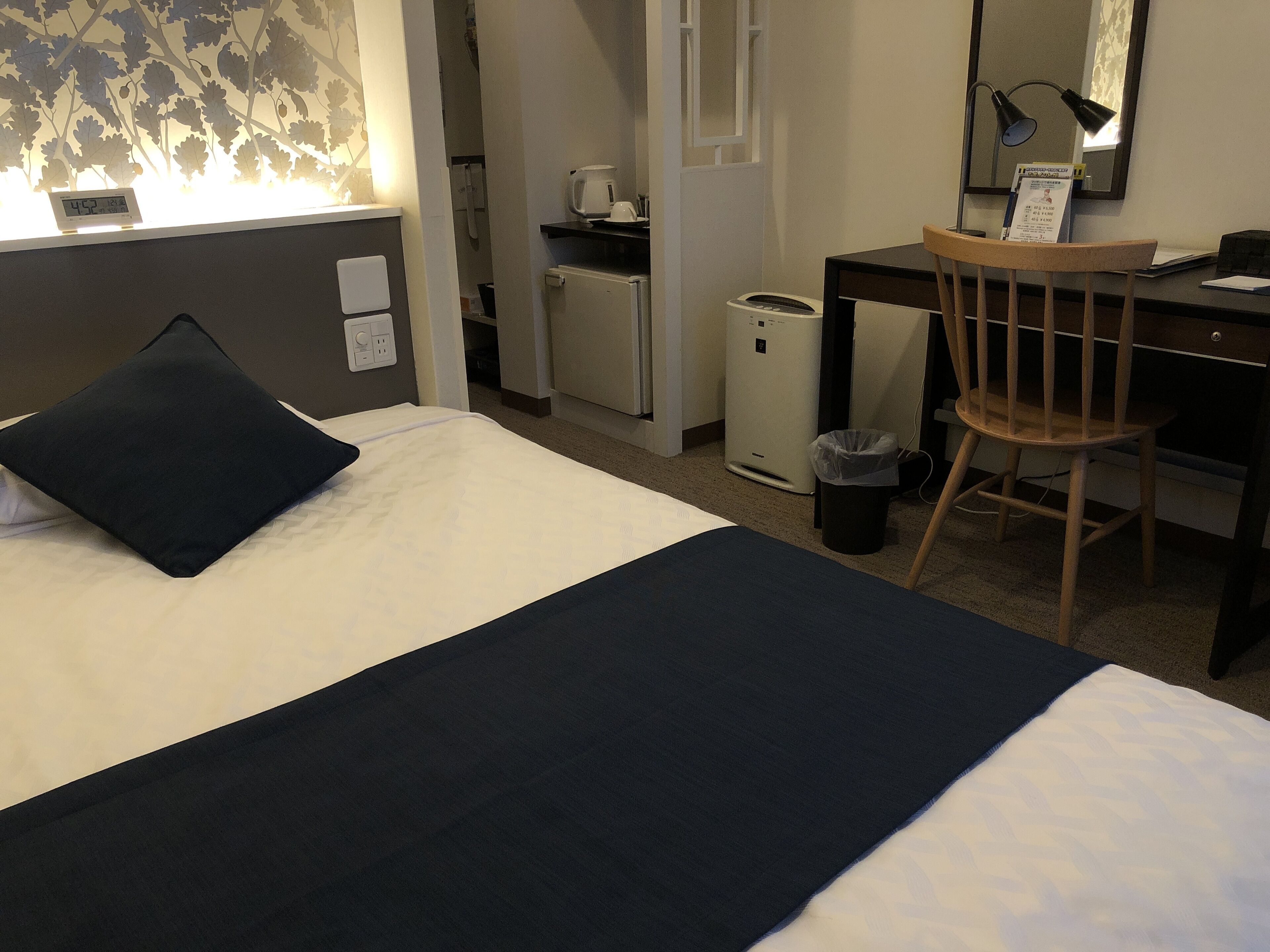 A hotel room with a bed, a desk, a chair, a trash bin, a coffee machine, and a clock.