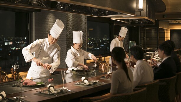 Un restaurante de alta categoría con chefs preparando comida en la barra, con vistas a un paisaje urbano nocturno.