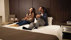 Una pareja sentada en una cama en una habitación de hotel con un panel de madera, una mesita de noche con lámpara y un reloj digital.