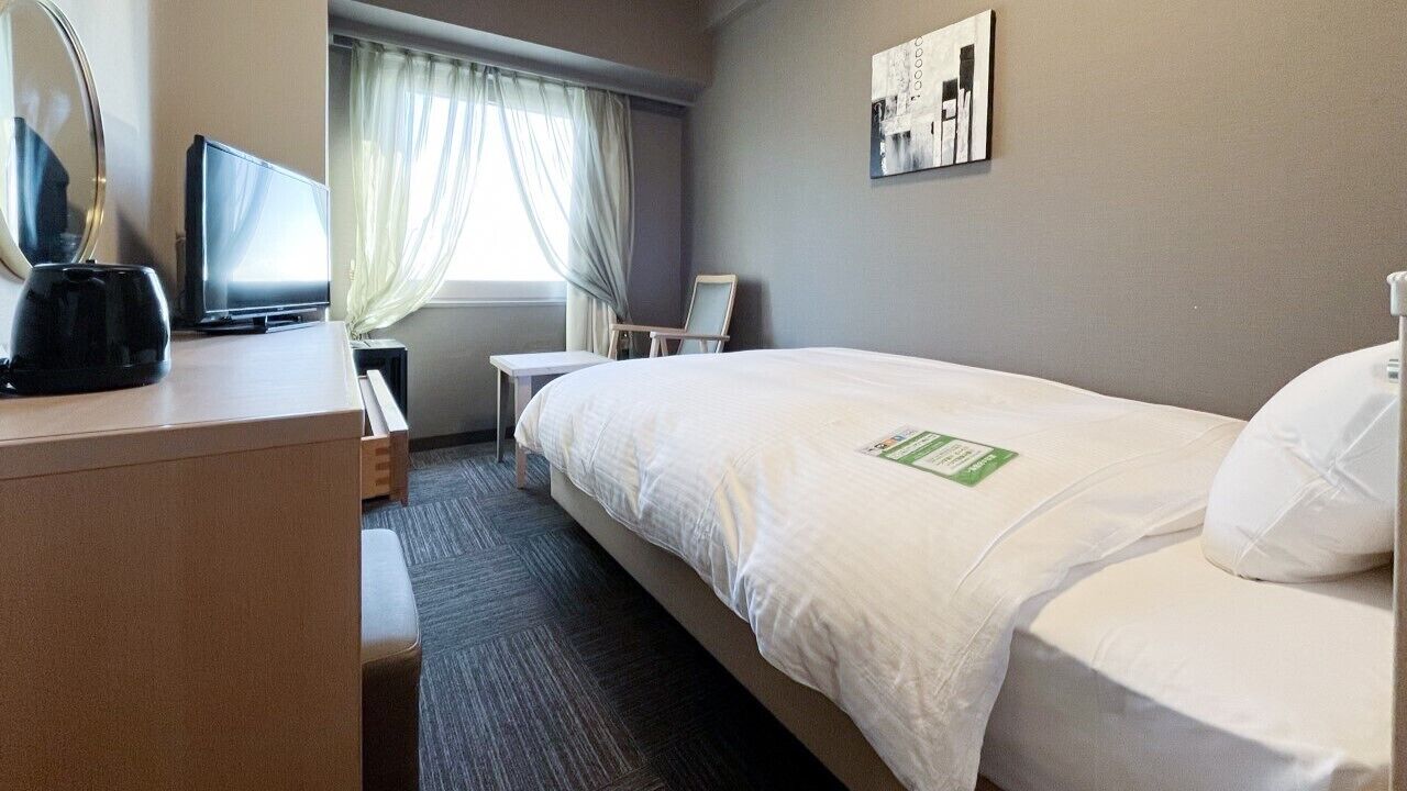 Une chambre d’hôtel avec un lit, un bureau avec un ordinateur, une chaise, une fenêtre avec des rideaux et un tableau au mur.