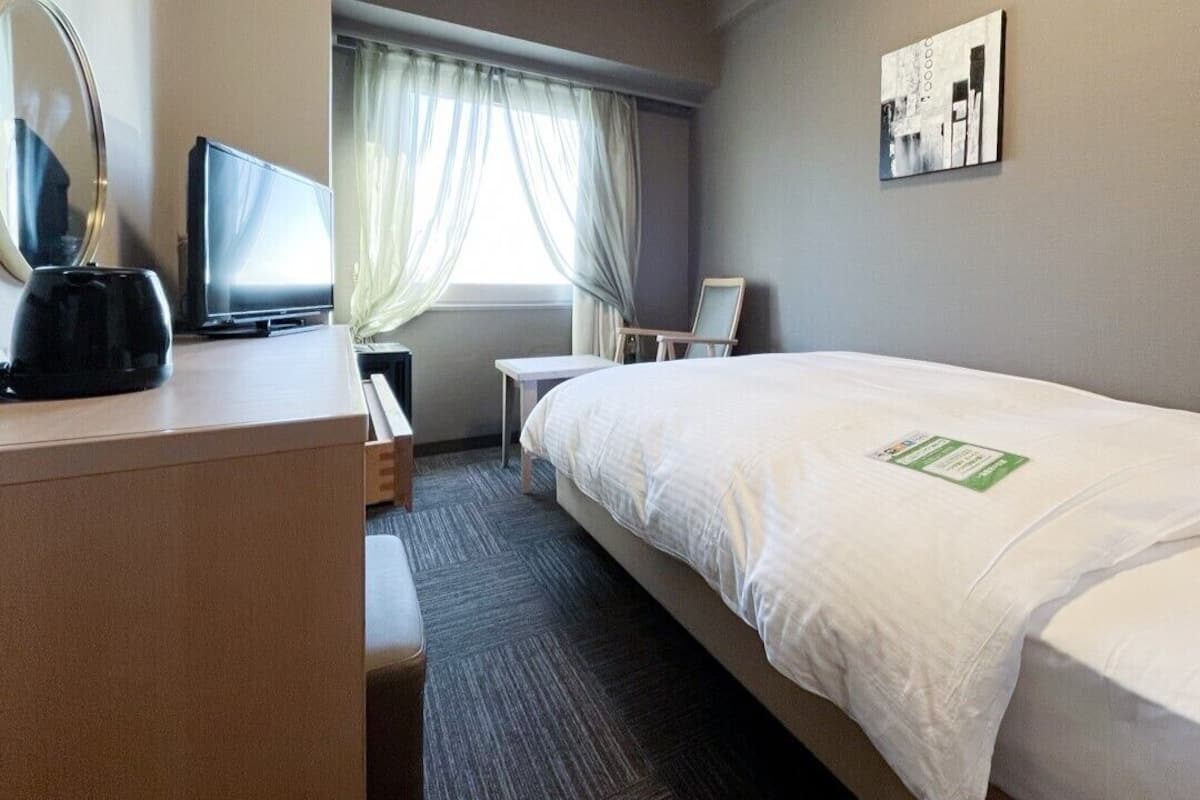 Una camera d'albergo con un letto, una scrivania con un computer, una sedia, una finestra con tende e un quadro appeso al muro.
