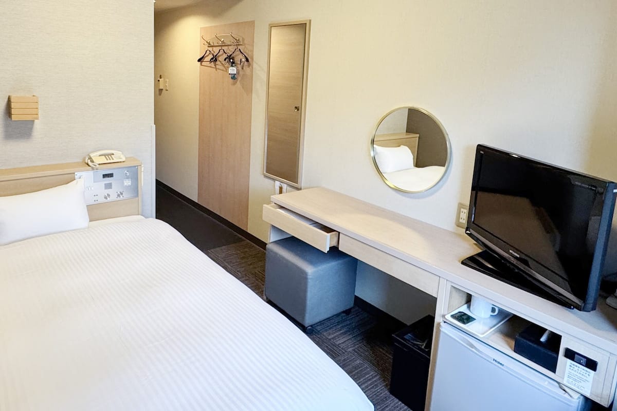 Camera d'albergo con un letto, telefono, televisione, scrivania, specchio e appendiabiti con grucce.