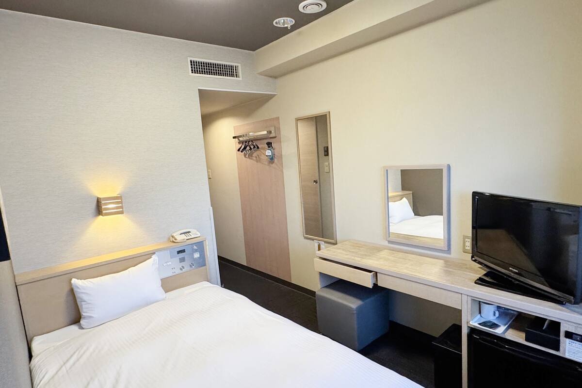 Una camera d'albergo con un letto, una scrivania con televisore, uno specchio, un telefono e un piccolo mobiletto.