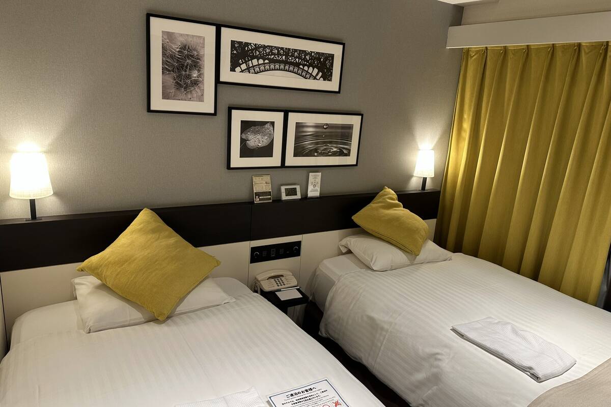 Habitación de hotel con dos camas, cada una con sábanas blancas y una almohada amarilla. Hay cuadros enmarcados en la pared sobre las camas, un teléfono en la mesita de noche y una ventana con cortinas amarillas.