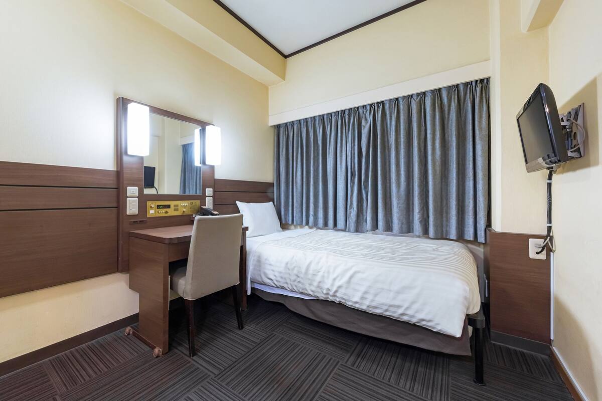 Una habitación de hotel con cama, escritorio con silla, espejo, televisor y ventana con cortinas.