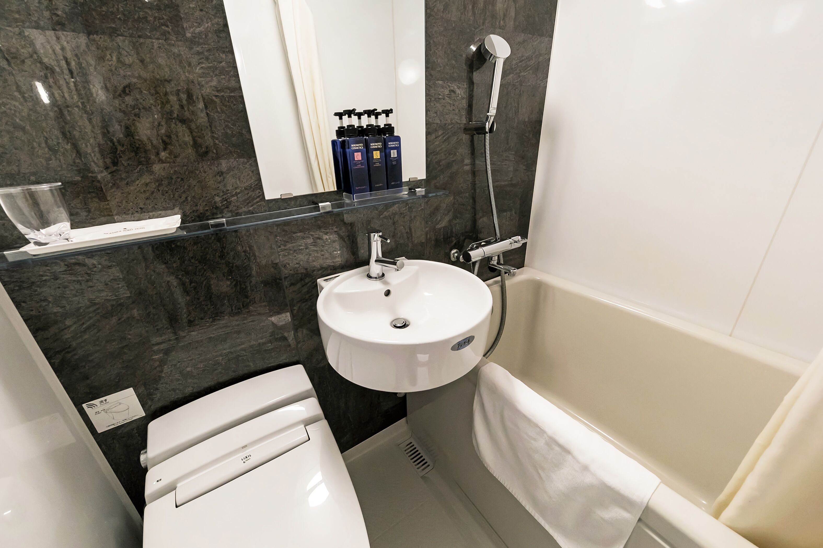 A compact bathroom with a white sink, a toilet, a bathtub, and a shower. There are toiletries on a shelf above the sink.