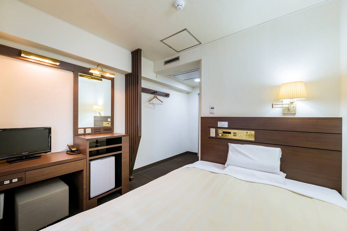 Habitación de hotel con cama, escritorio con televisor, espejo, lámpara y un refrigerador pequeño.