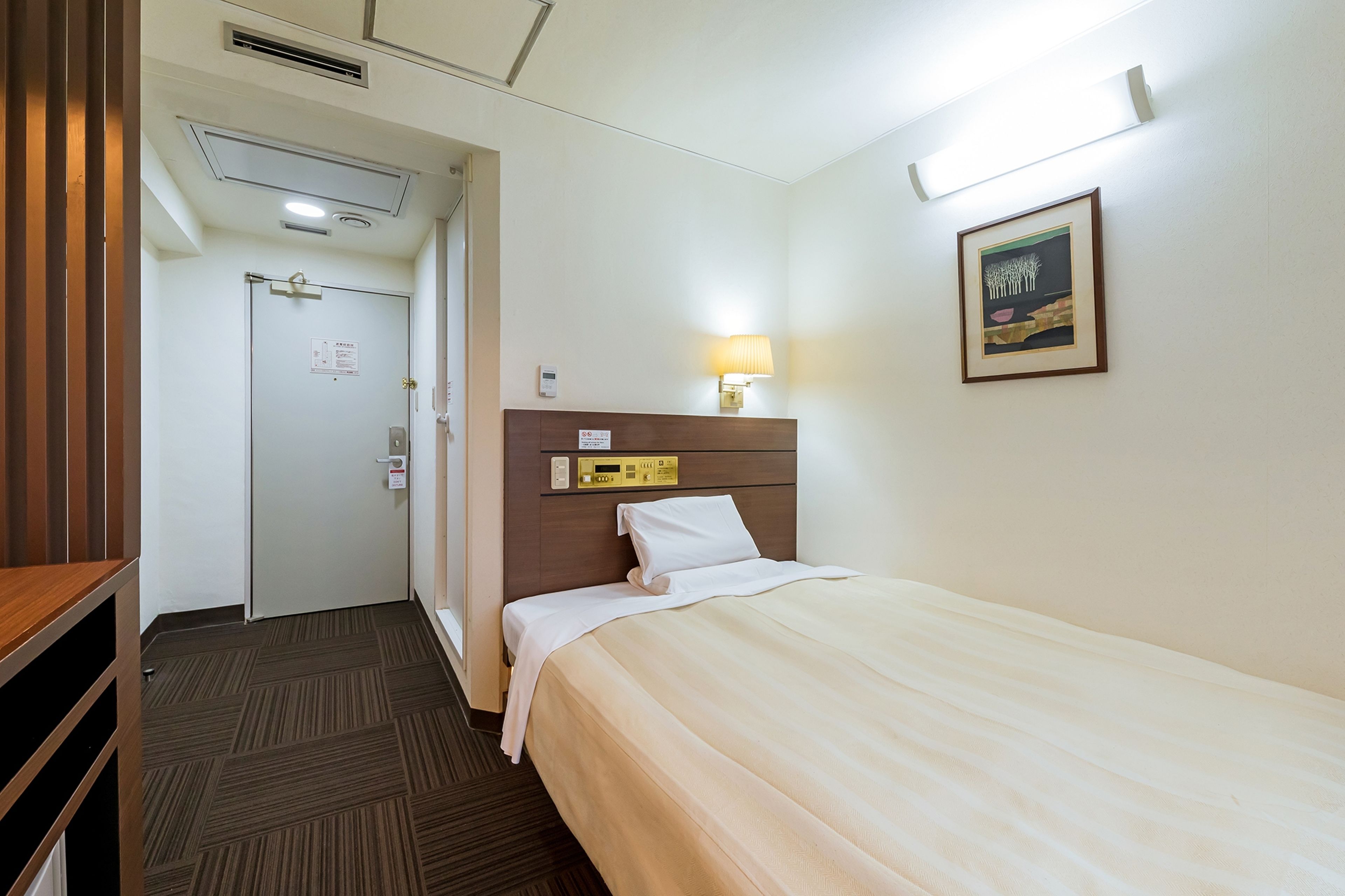 A hotel room with a bed, a bedside table, a lamp, a framed picture on the wall, and a door leading to another room.