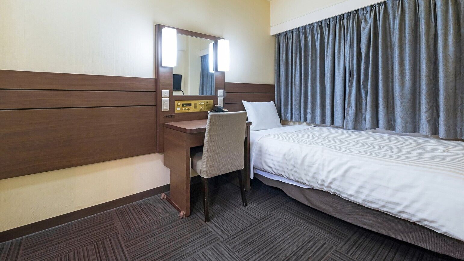 A hotel room with a bed, a desk with a chair, a mirror, and a control panel.