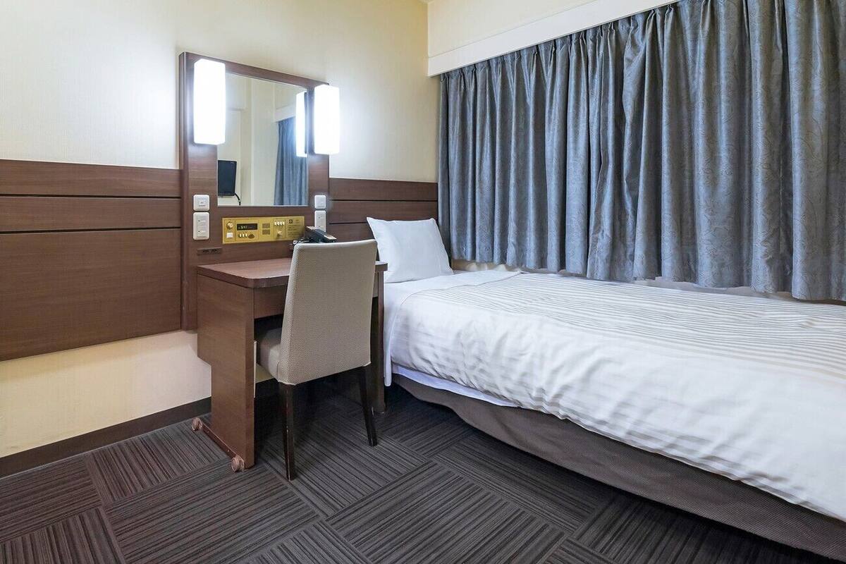 Una habitación de hotel con una cama, un escritorio con una silla, un espejo y un panel de control.