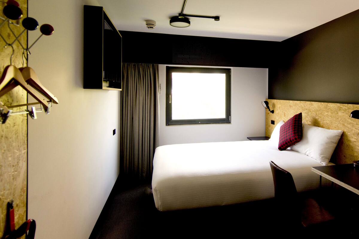 A hotel room with a bed, a desk, a chair, a hanging coat rack, and a window with curtains.