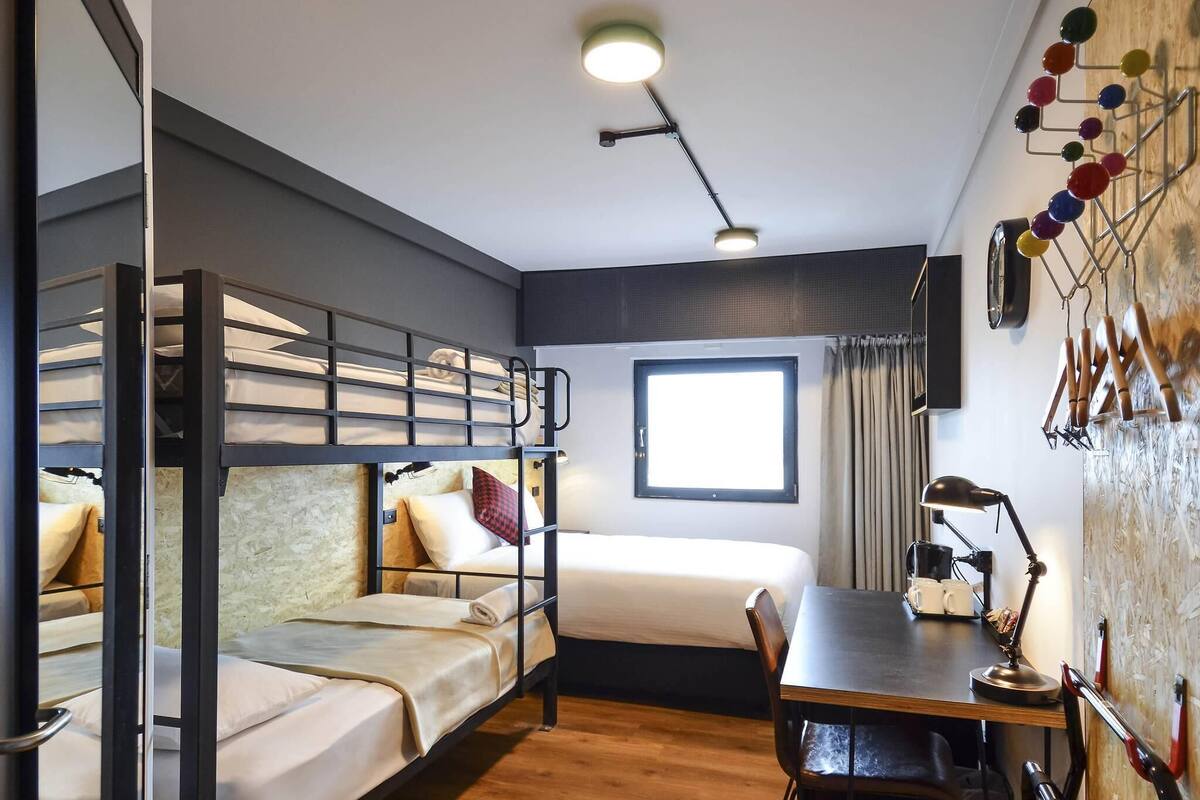 A dormitory room with bunk beds, a desk, a chair, a lamp, and a window.
