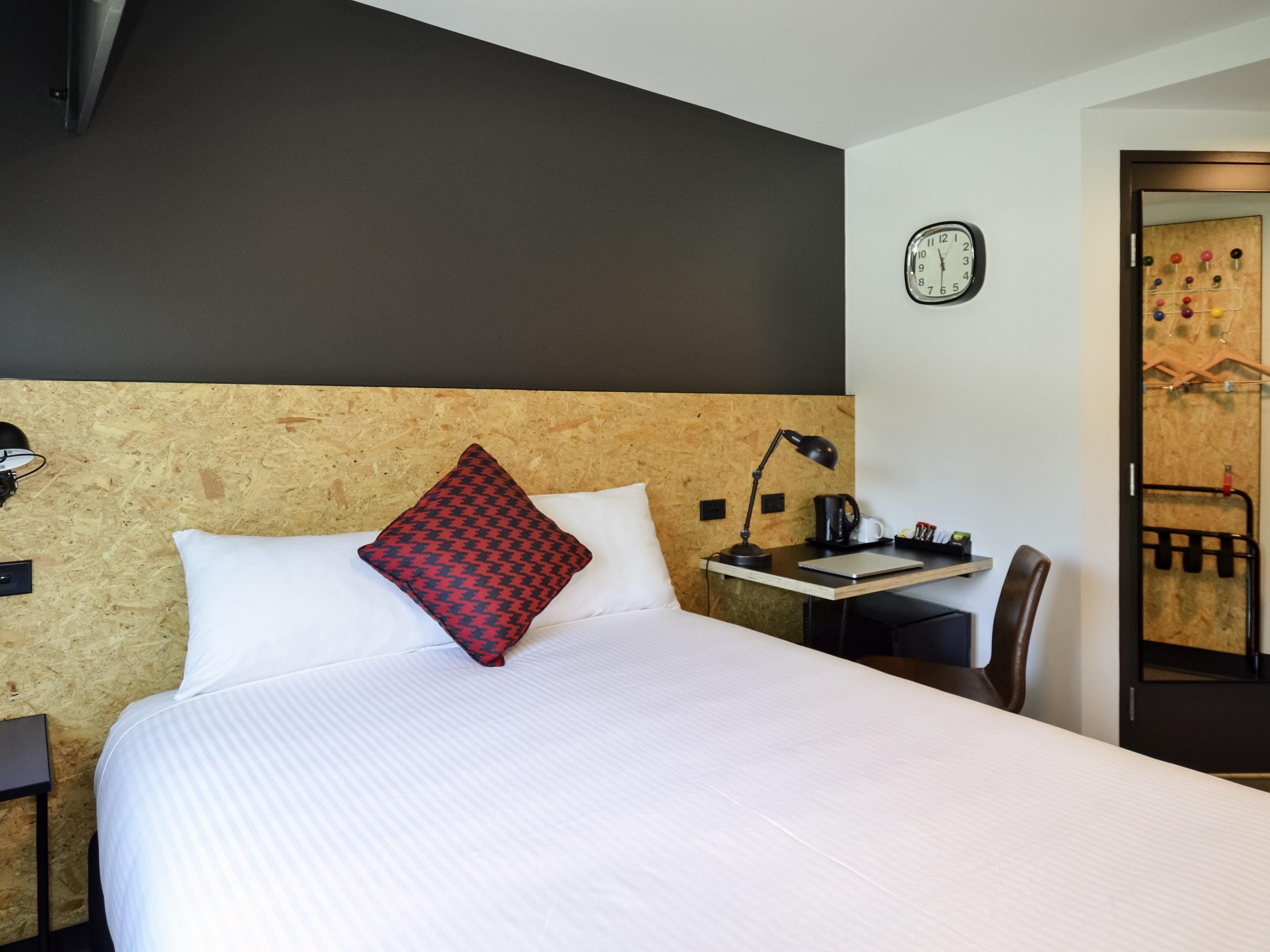 A hotel room with a bed, a desk with a lamp, a clock on the wall, and a door with a coat rack.