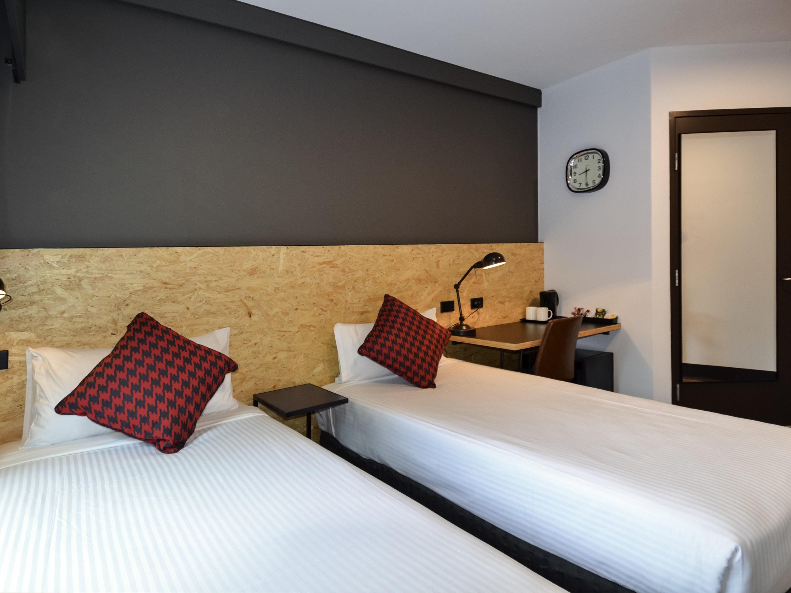 A hotel room with two beds, a desk, a chair, a clock, and a door.