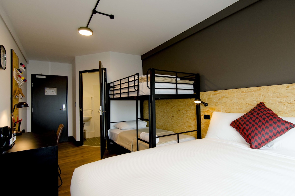 A hotel room with a bunk bed, a desk with a lamp, and a bed with a pillow.