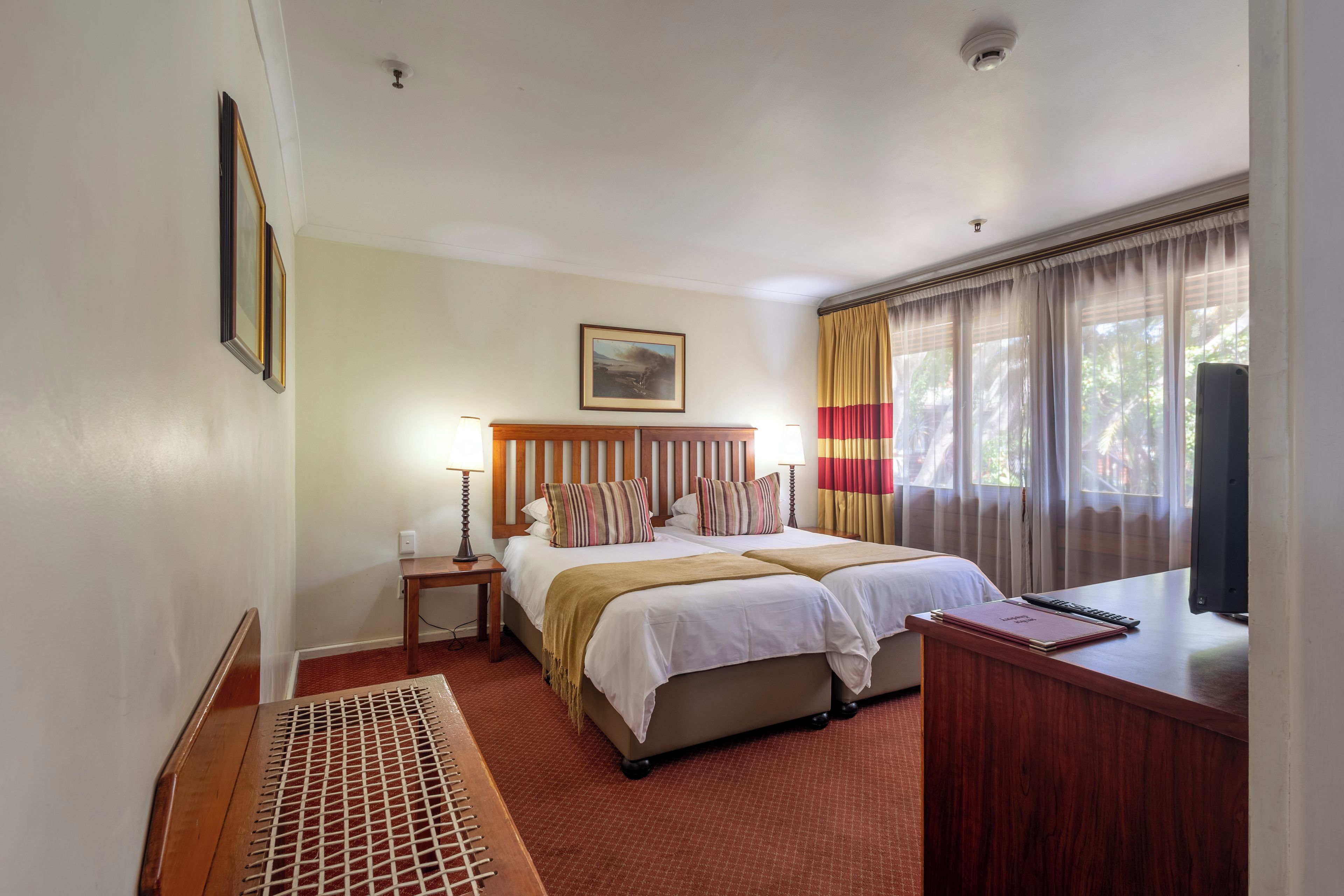 Habitación de hotel con dos camas, un escritorio, una silla, un radiador y una ventana con cortinas.