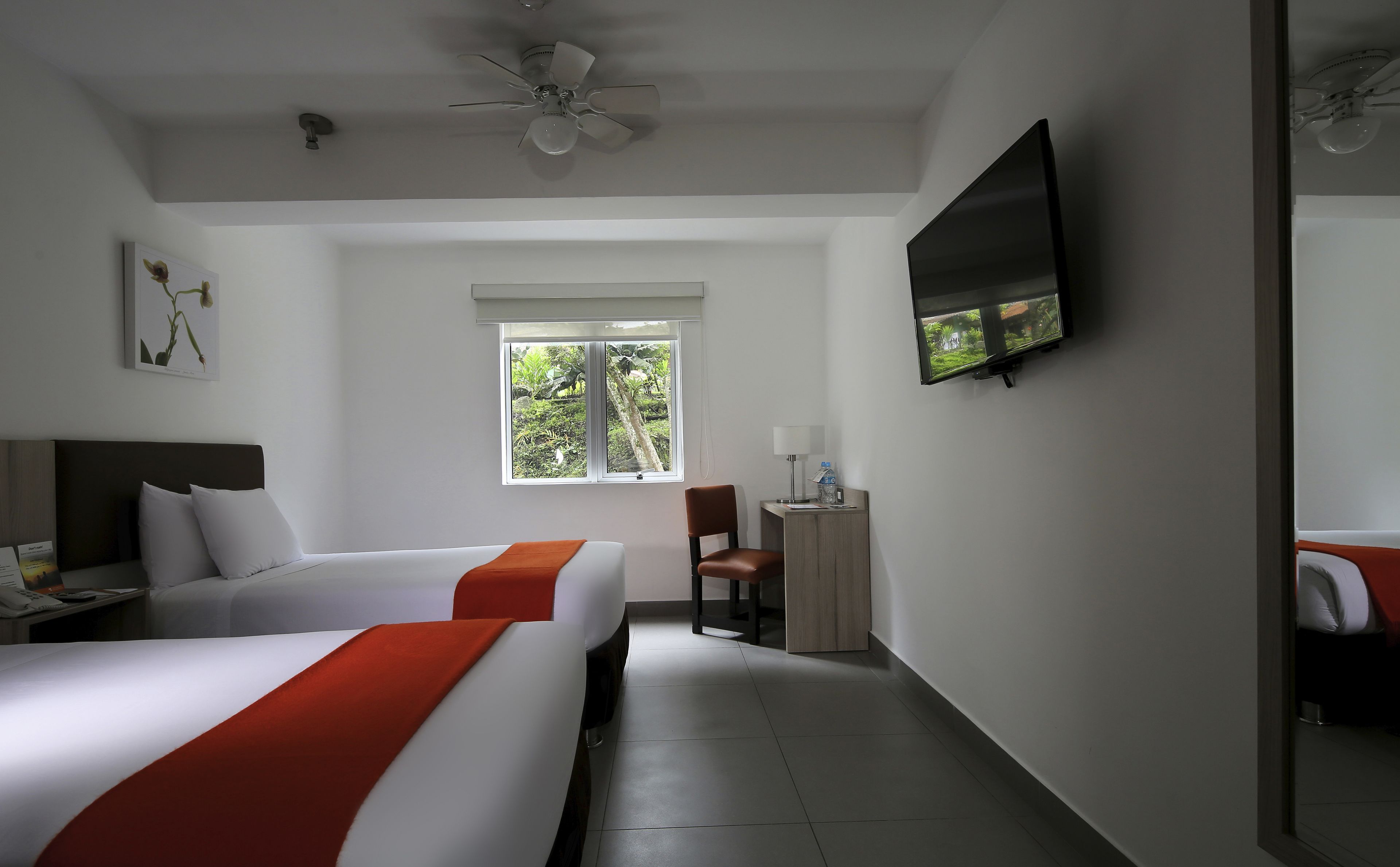 A hotel room with two beds, a TV, a window with a view of greenery, a ceiling fan, and a desk with a lamp.