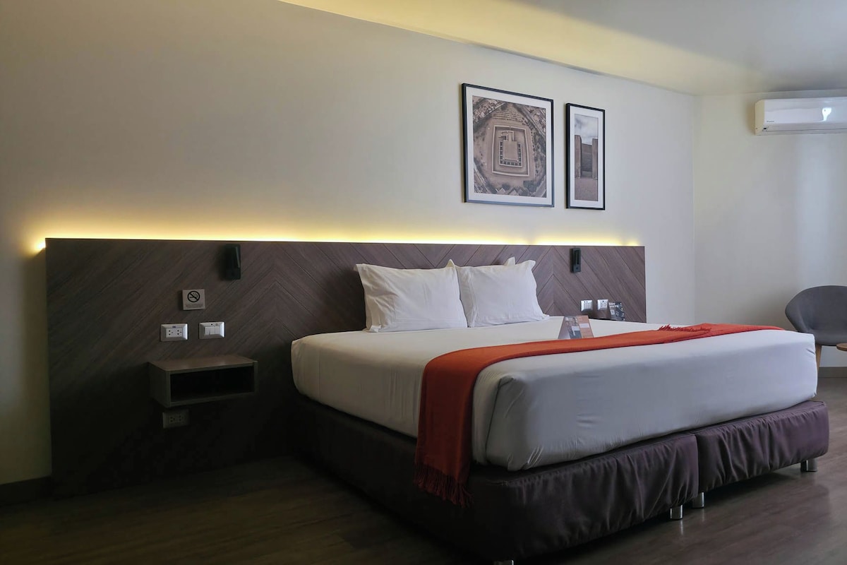 Habitación de hotel con una cama grande, mesitas de noche, una silla y una obra de arte en la pared.