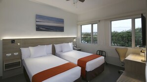 Hotelkamer met twee bedden, een bureau, een stoel, een plafondventilator, een groot raam en een schilderij aan de muur.
