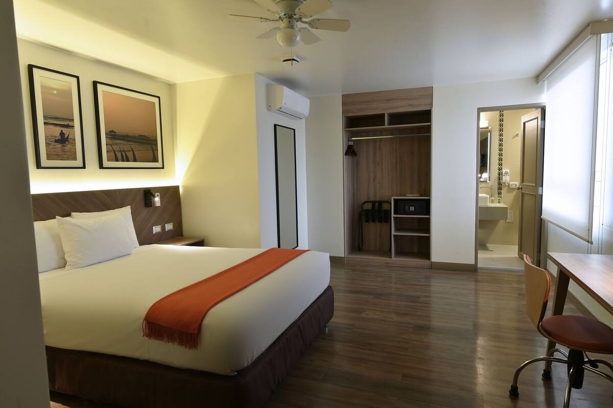 Une chambre d’hôtel avec un lit, un bureau, une chaise, une armoire et une salle de bain visible à travers une porte ouverte.