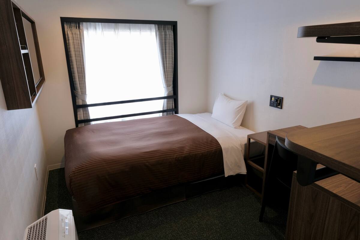 A hotel room with a bed, a desk, a chair, a window with curtains, and a wall-mounted shelf.