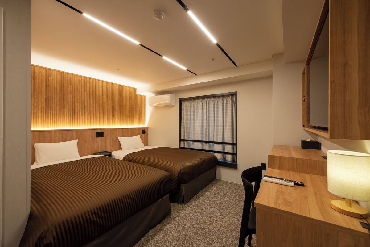 A hotel room with two beds, a desk, a chair, a lamp, a window with curtains, and a wooden headboard.