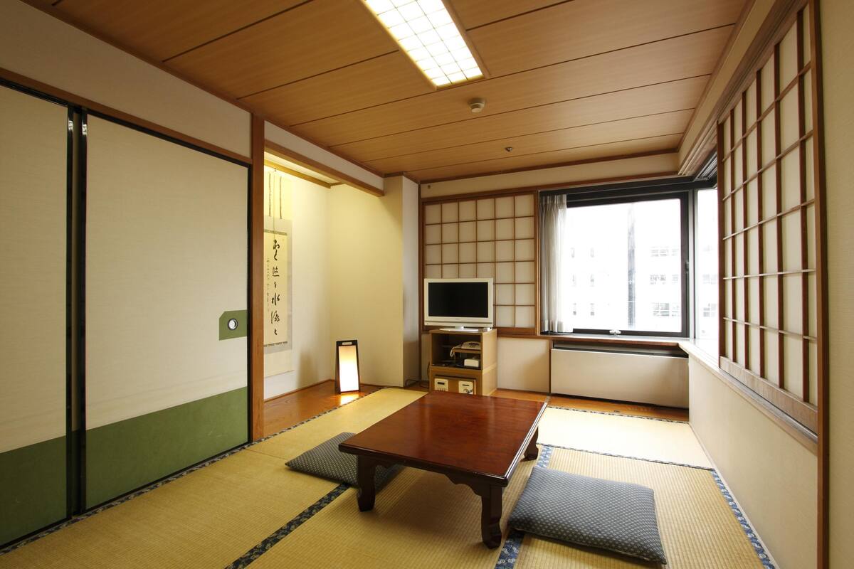 A traditional Japanese room with tatami flooring, a sliding door, a television, and a window with a view of buildings.