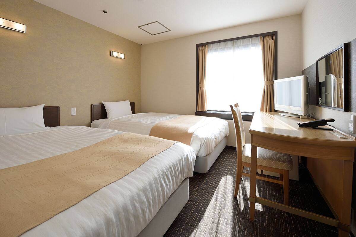 A hotel room with two beds, a desk, a chair, a television, and a large window with curtains.